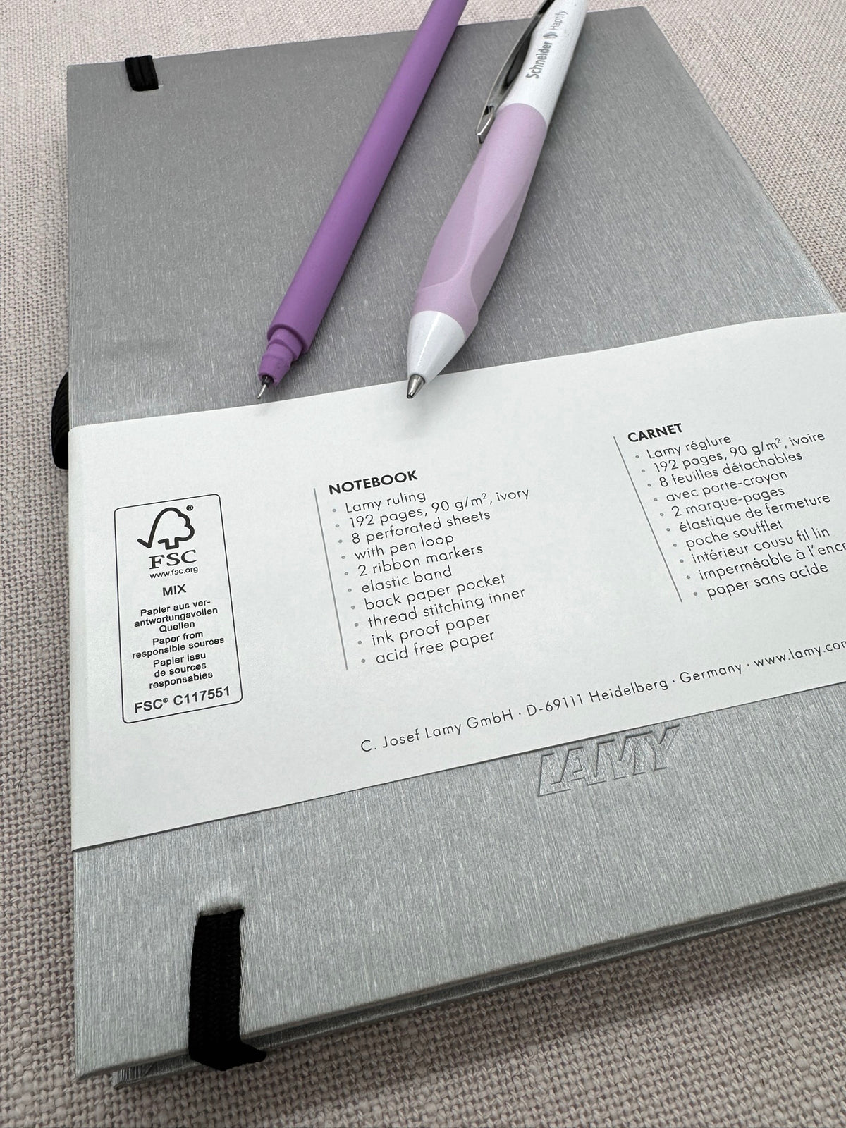 Auf der Rückseite des Notizbuches sieht man ein dezentes Lamy Logo ebenfalls in Silber. Auf dem Umschlag sind alle Details abgebildet: 192 Seiten, 90 g Papier, PPen Loop, 2 Stofflesezeichen, hinten eine Tasche, Papier kann auch mit Füller beschrieben werden. Auf dem Notizbuch liegen zwei Stifte nebeneinander: ein Fineliner in Lila und und Kugelschreiber in Weiß-Flieder. 