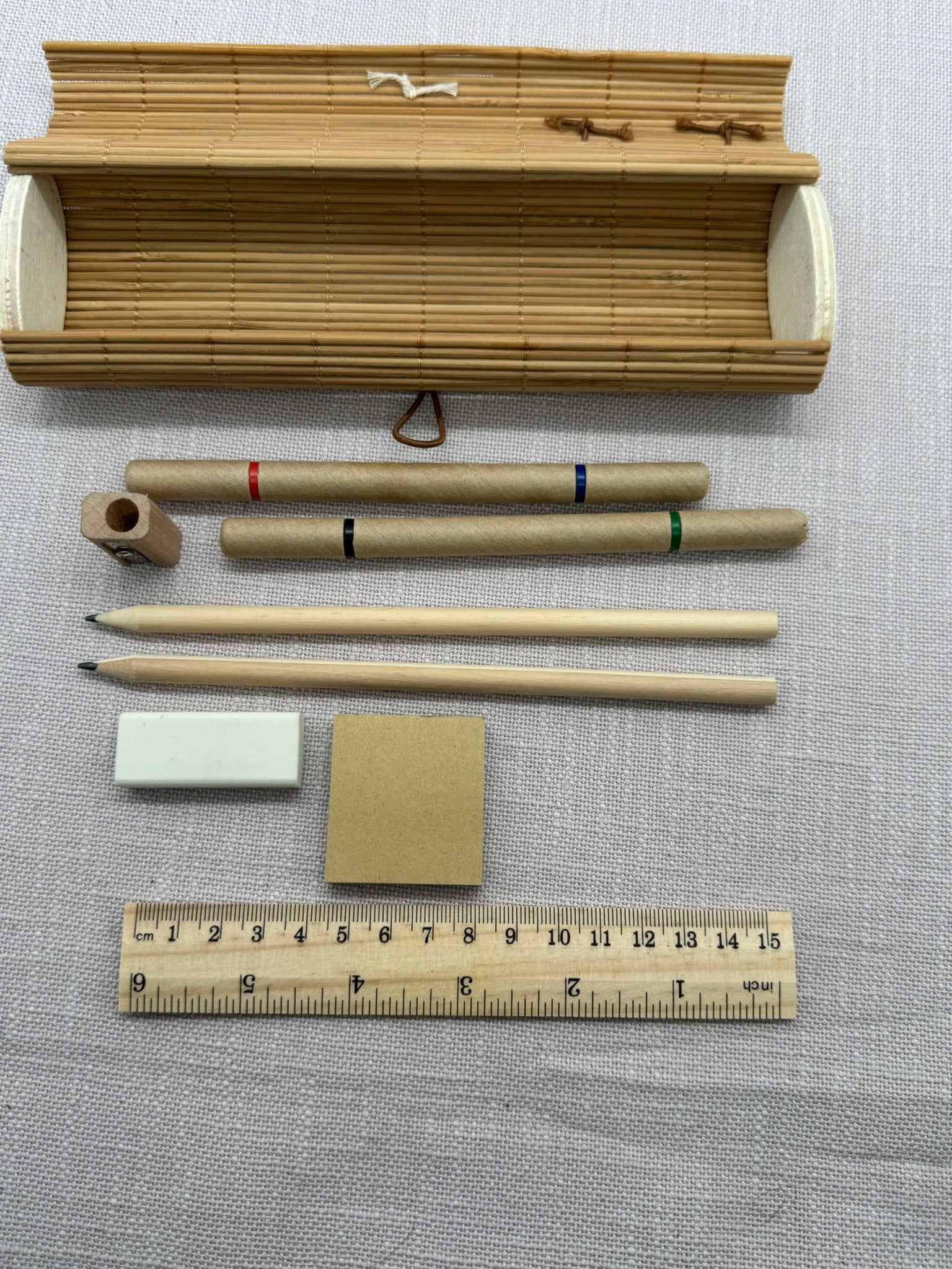 Alle Schreibwaren im Set ausgepackt auf hellem Hintergrund: Etui aus kleinen Holzstäbchen, zwei Stifte mit zwei Spitzen, ein Bleistiftanspiter aus Holz, zwei Bleistifte aus unbehandeltem Holz, ein heller Radierer, kleine Haftnotizen in beige, ein Lineal aus Holz, 15 cm lang. 