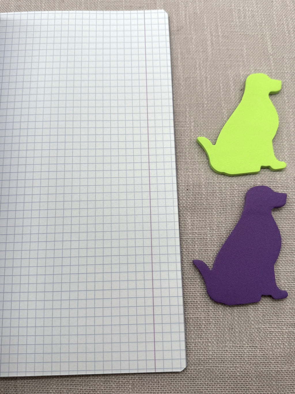 Aufgeschlagenes Heft neben Haftnotizen in Hundeform in Neonrün und Lila: man sieht eine karierte Seite mit Rand. 