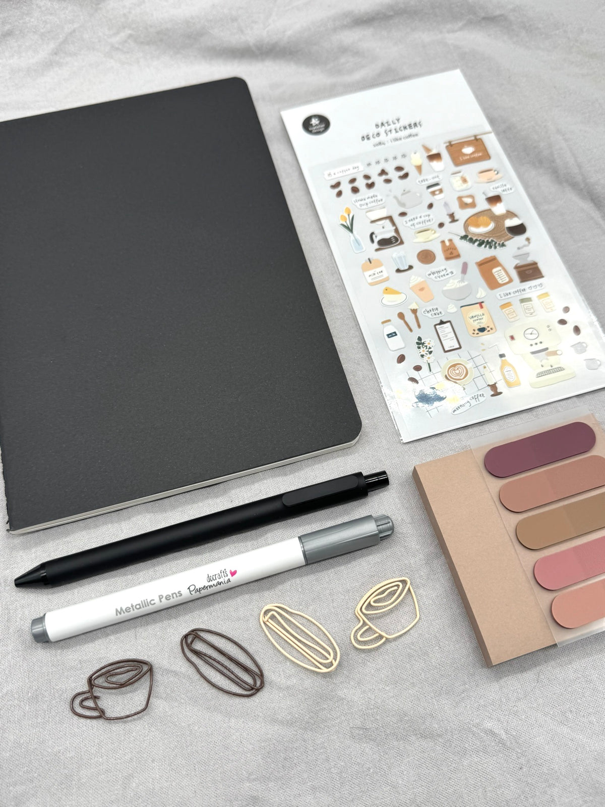 In dem Foto ist eine Übersicht über den Inhalt des Schreibsets in Schwarz abgebildet. Das Heft und der Gelstift sind Schwarz, der Metallic-Pen dagegen Silber. 