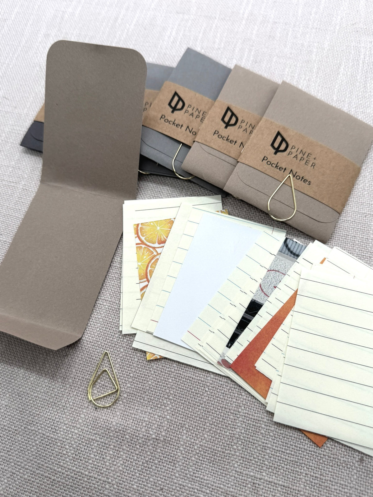 Man sieht ein auseinandergebautes Heftchen, bestehend aus 25 Notizzetteln aus unterschiedlichen Papieren, einer Verpackung aus Kraftpapier und einer goldenen Büroklammer. 