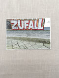 Ein zwischen Berlin-Mitte und Friedrichshain aufgenommenes Bild von Street Art. "Zufall" steht in großen silbernen Buchstaben auf einer Häuserwand.