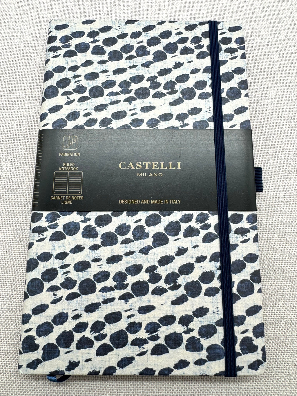 Das Notizbuch im Set ist von der Firma Castelli und wurde in Italien hergestellt. Es hat ein helles Cover mit unregelmäßigen Punkten in Dunkelblau. Das Cover ist aus einem Stoff. Dazu hat es ein Gummiband und eine Stiftschlaufe in Schwarz. Das Etikett sagt, es sei liniert. 