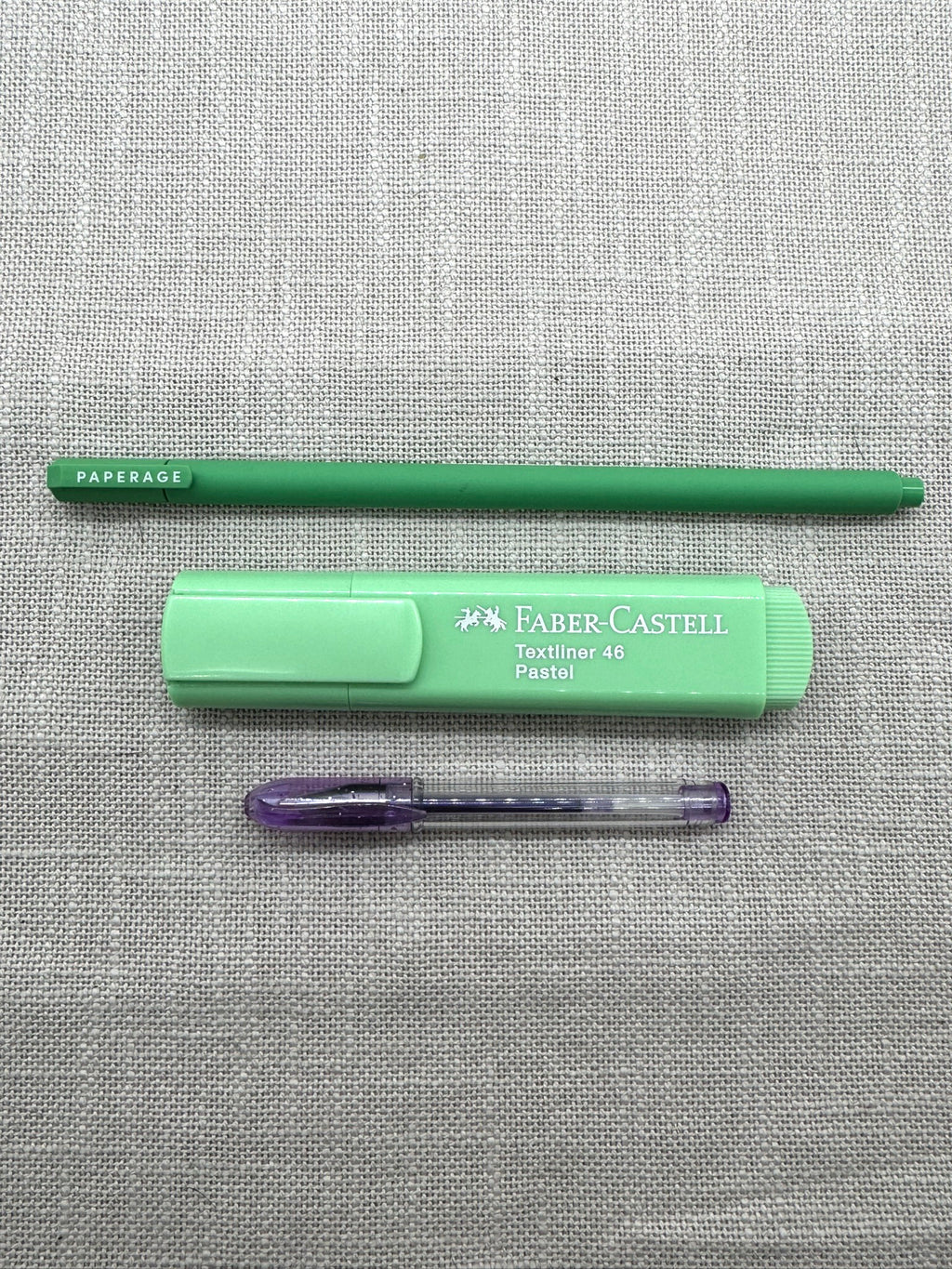 Diese Schreibwaren sind im Set enthalten: ein Paperage Fineliner in Grün, ein Textmarker von Faber-Castell in mintgrün, ein kleiner Gelstift in Lila. Die Stifte liegen nebeneinander auf einem Leinenstoff. 