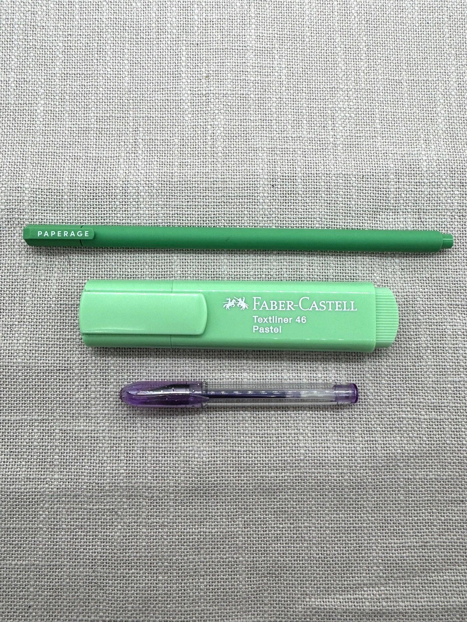 Diese Schreibwaren sind im Set enthalten: ein Paperage Fineliner in Grün, ein Textmarker von Faber-Castell in mintgrün, ein kleiner Gelstift in Lila. Die Stifte liegen nebeneinander auf einem Leinenstoff. 