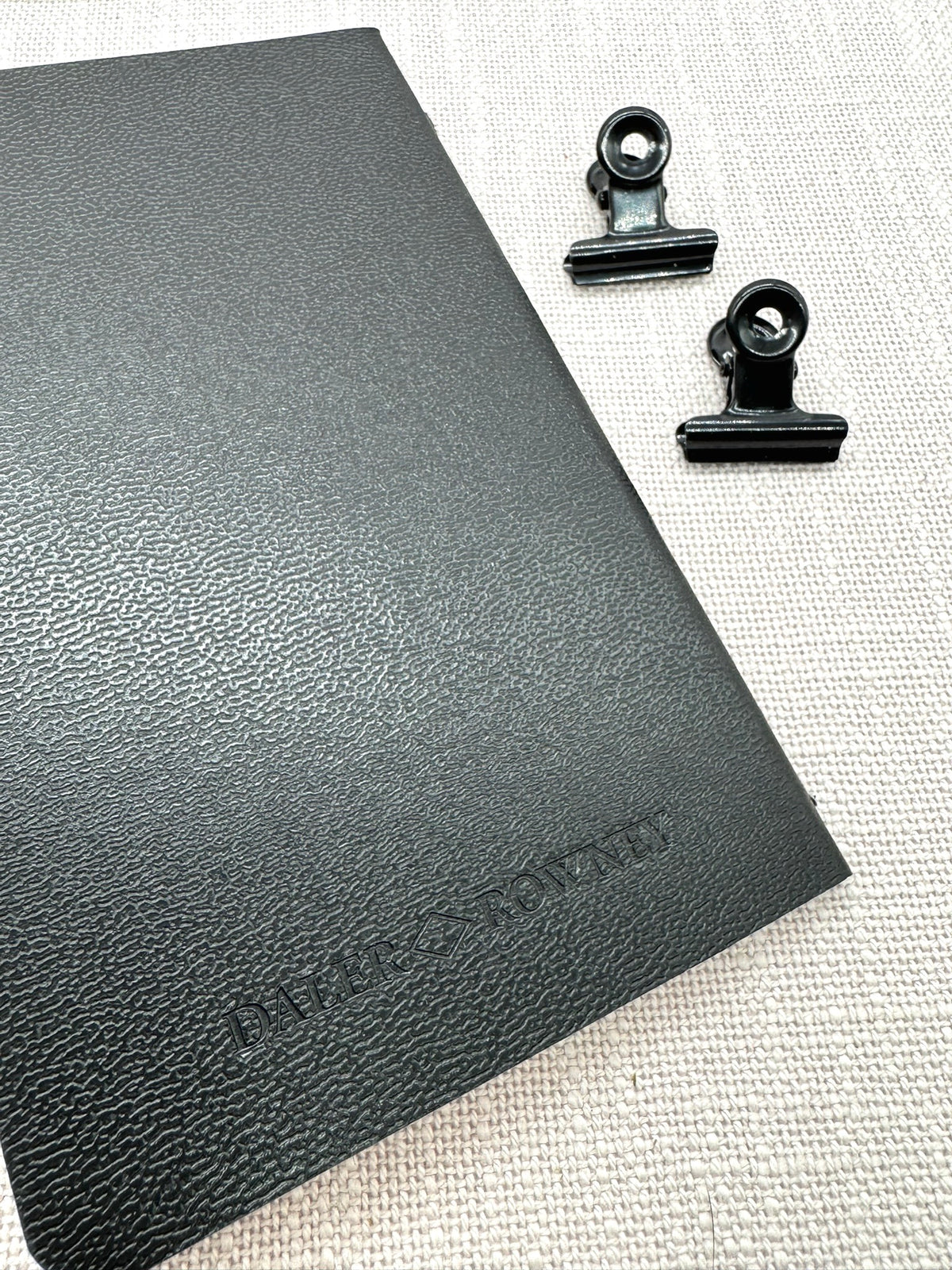 Die Rückseite des Notizbuchs: Das Cover ist schwarz, man sieht ein sehr dezentes Logo ebenfalls in Schwarz. Daneben liegen zwei schwarze Papierklemmer. 