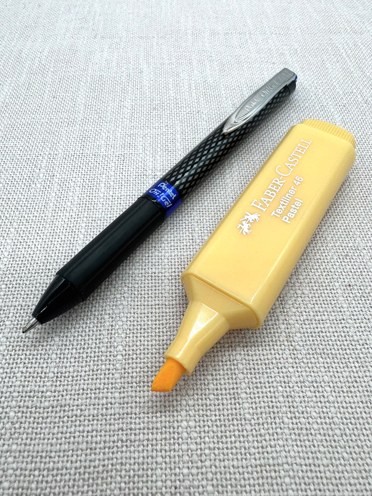 Ein Gel Pen von Pentel liegt neben einem Textmarker von Faber-Castell. Der Stift ist Schwarz mit einem silbernen Clip, der Marker ist Pfirsich-Orange. Man sieht eine Nahaufnahme der Spitzen. 