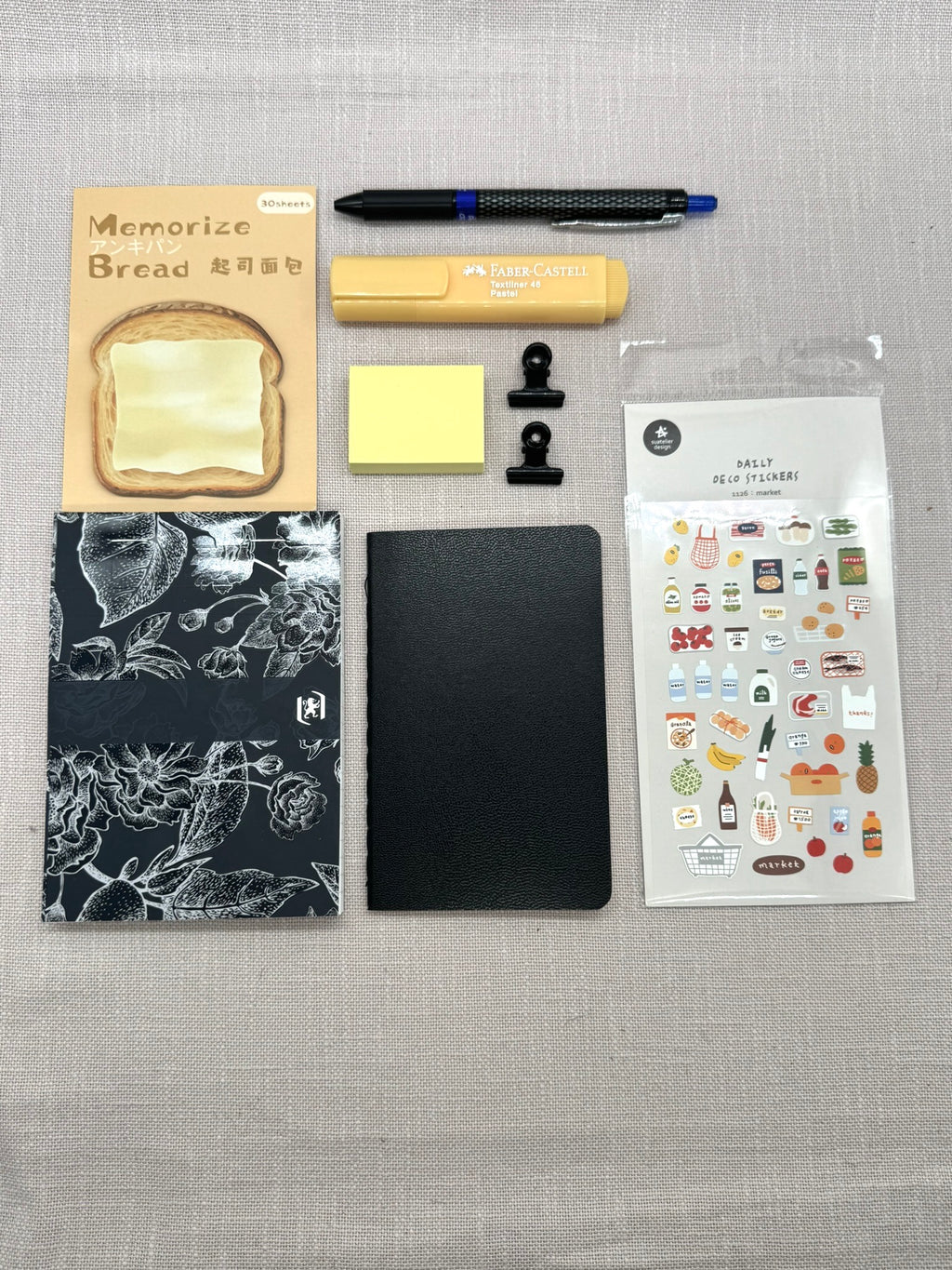 Das Schreibwaren-Set von oben fotografiert auf einem hellen Leinentuch: Notizblock, daneben ein Notizbuch in A6, koreanische Mini-Sticker, Haftnotizen einmal normal, einmal als Brotscheibe, Textmarker und Gelstift, darunter Metallklammern in Schwarz