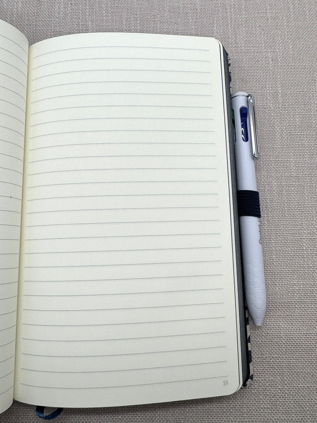 Das Heft liegt aufgeschlagen auf einem hellen Leinenhintergrund. Man sieht eine linierte Seite mit Seitenzahlen. Der Pentel-Kulli steckt in der Stiftschlaufe. 