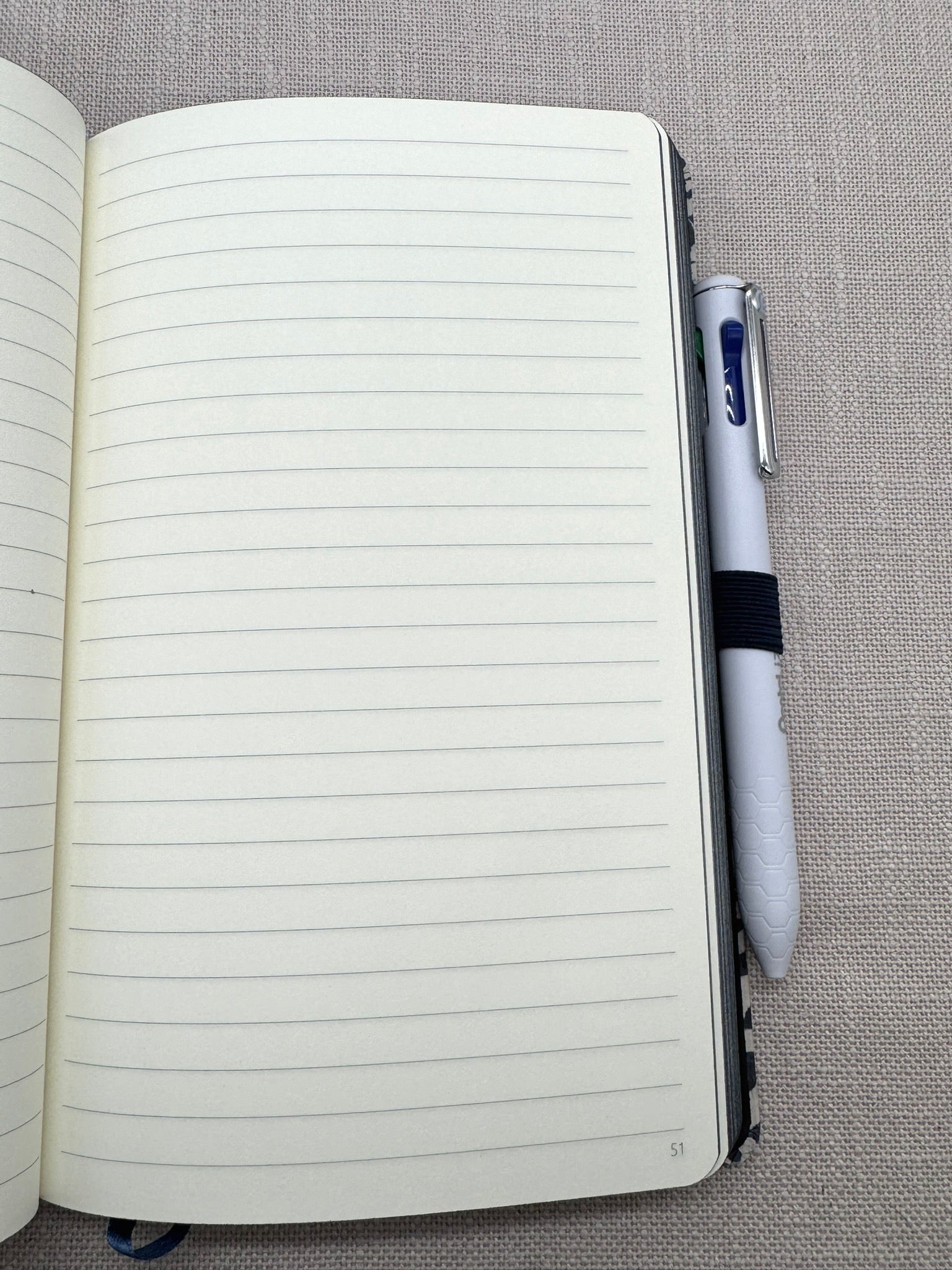 Das Heft liegt aufgeschlagen auf einem hellen Leinenhintergrund. Man sieht eine linierte Seite mit Seitenzahlen. Der Pentel-Kulli steckt in der Stiftschlaufe. 