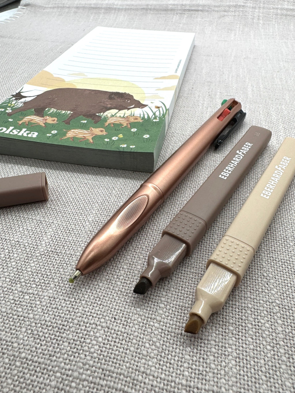 Im Vordergrund sieht man die Spitzen der Schreibwaren: Der 4-Farben-Kugelschreiber liegt neben dem braunen Marker, daneben der Marker in beige. 
