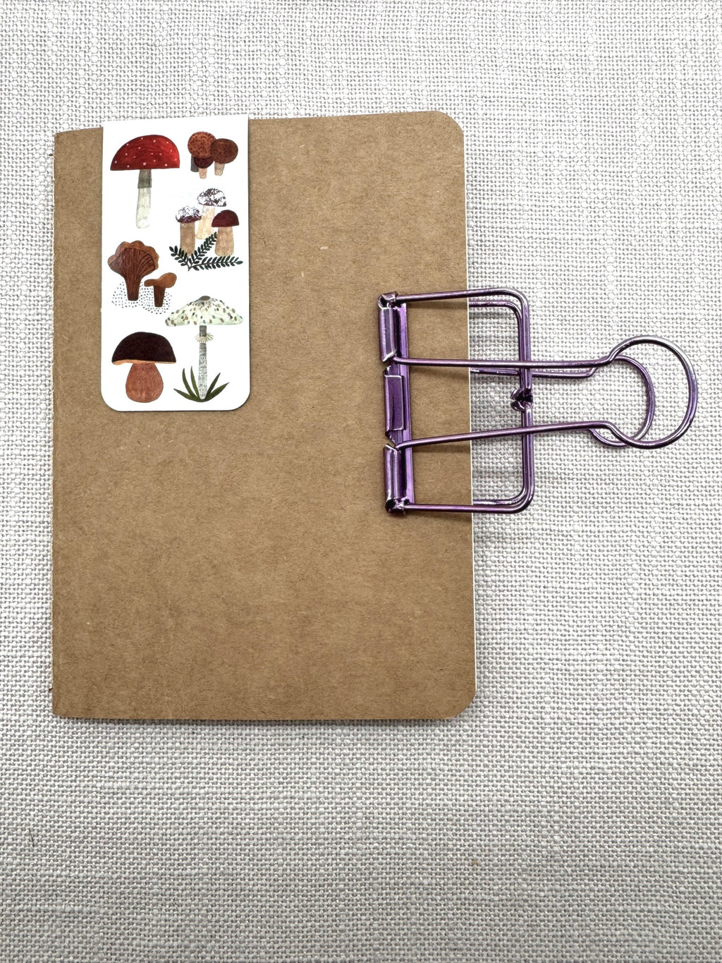 Ein Notizbuch in A6 mit Cover aus Kraftpapier. Oben ist ein magnetisches Lesezeichen mit Pilzen eingesteckt. An der Seite klemmt eine Briefklemme in metallic-lila.