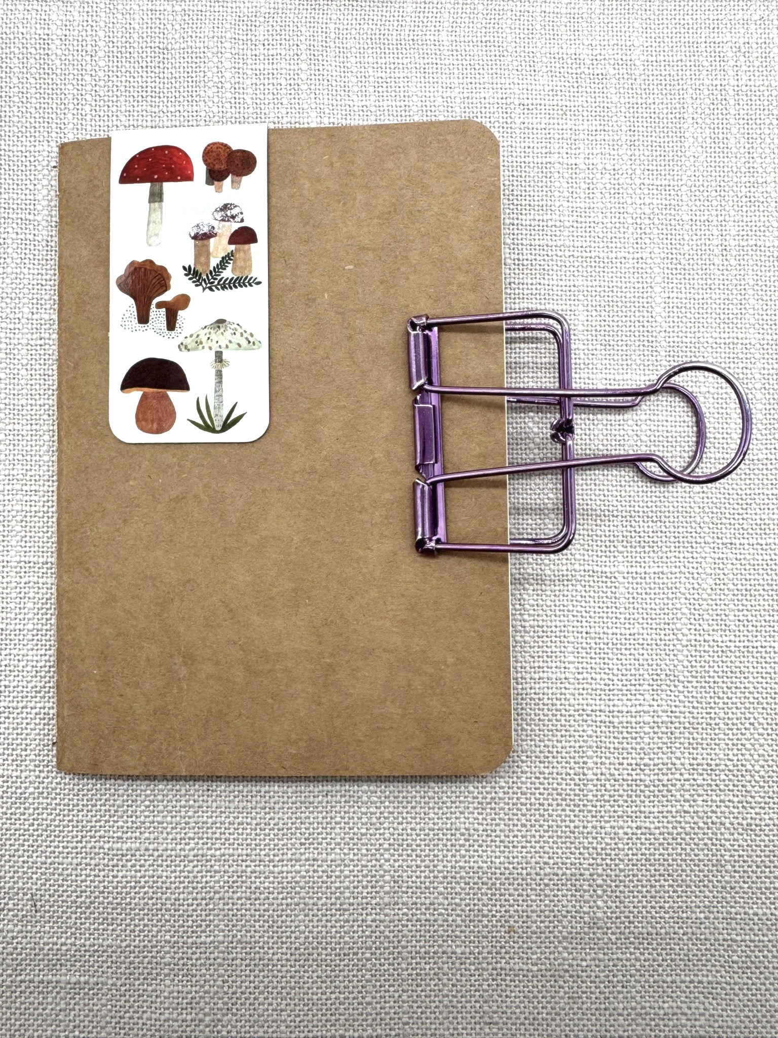 Ein Notizbuch in A6 mit Cover aus Kraftpapier. Oben ist ein magnetisches Lesezeichen mit Pilzen eingesteckt. An der Seite klemmt eine Briefklemme in metallic-lila.