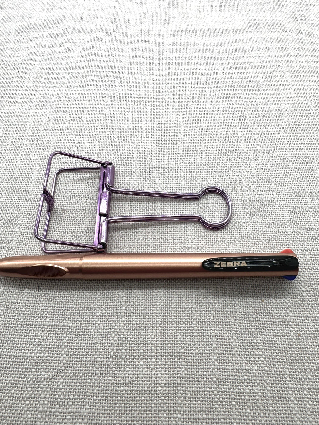 Nahaufnahme: Oben liegt eine Foldback-Klammer in metallic-lila, unten der 4-Farben-Kugelschreiber von Zebra