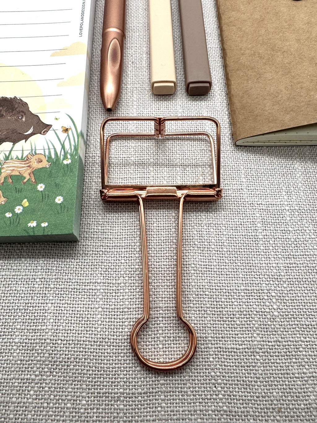 Im Vordergrund sieht man die Metallklammer in rosegold bzw. leich kupferfarben. Darüber liegen die Textmarker in beige und braun sowie der Kugelschreiber.