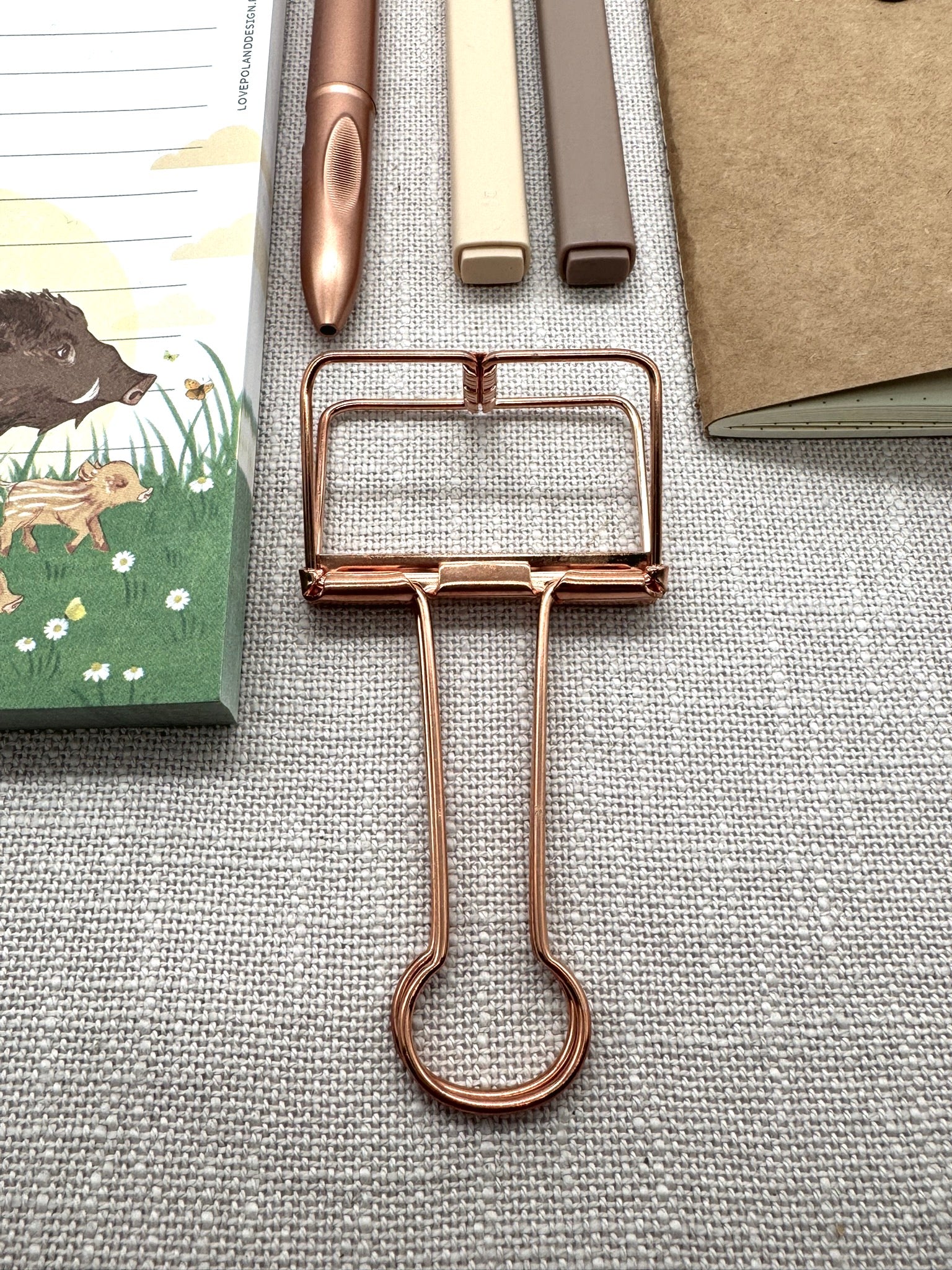 Im Vordergrund sieht man die Metallklammer in rosegold bzw. leich kupferfarben. Darüber liegen die Textmarker in beige und braun sowie der Kugelschreiber.