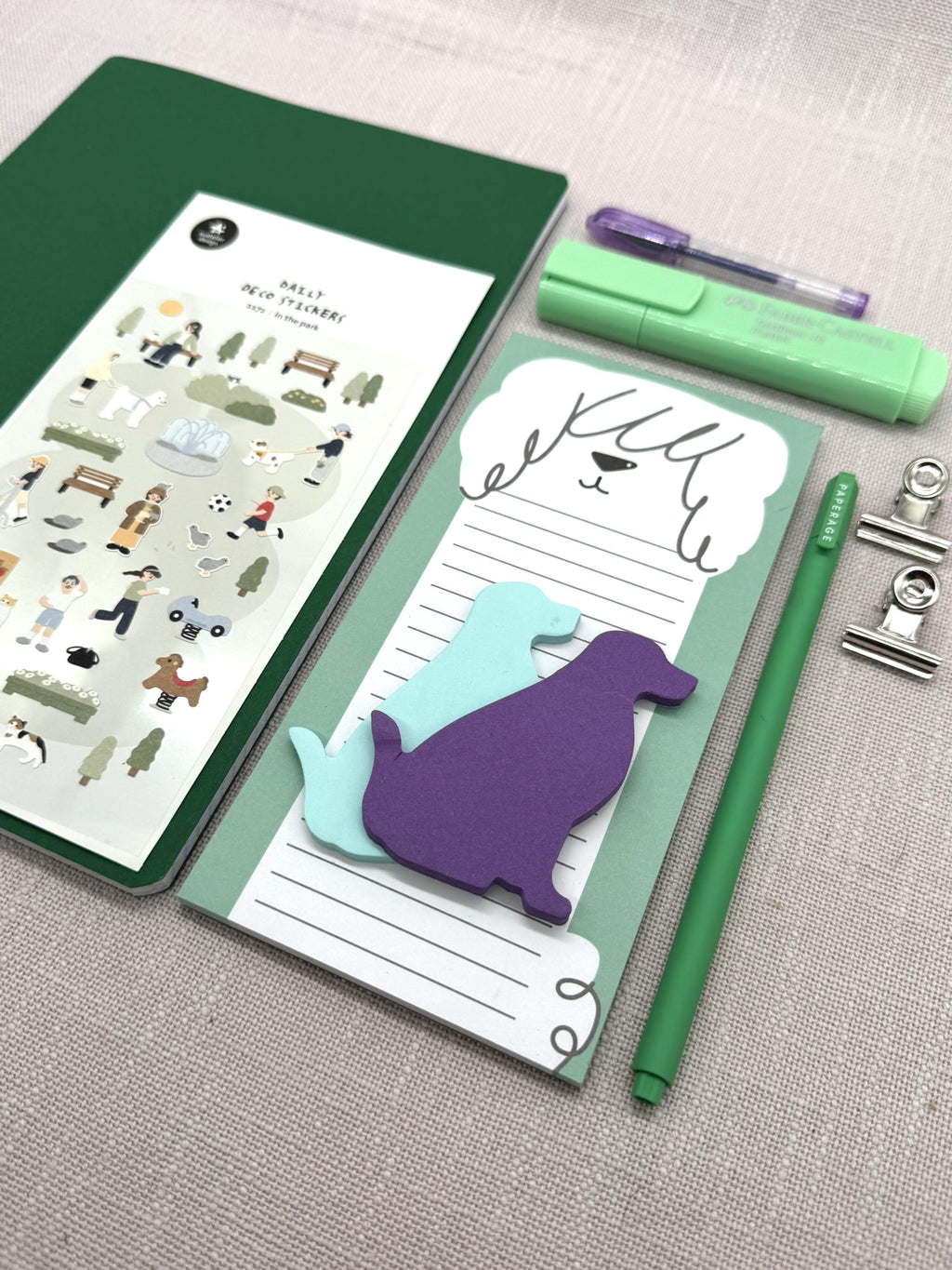 Ein Organisationsset mit Hundemotiv. Auf dem großen grünen Heft liegt ein Stickerbogen. Daneben eine To-Do-Liste mit Hundmotiv, darauf Post-Its in Hundeform. Ein schmaler Fineliner, ein Textmarker und ein kleiner Gelstift sind dabei. Sowie zwei kleine silberne Briefklammern. 