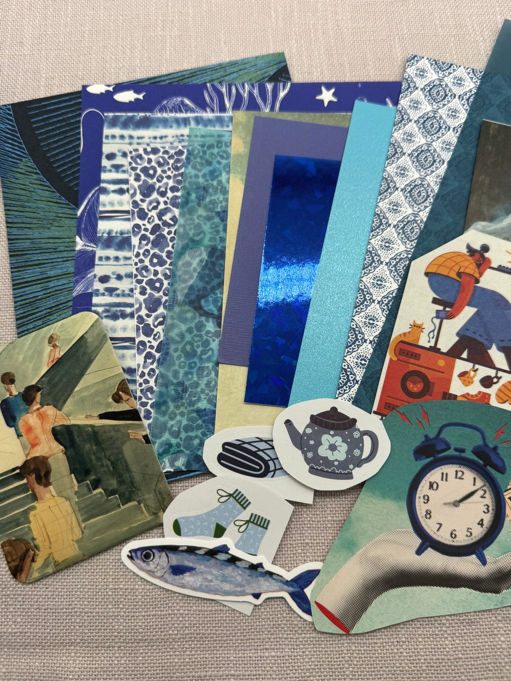 diverse blaue Papierausschnitte in verschiedenen Mustern, einfarbig, Transparentpapier, Glitzerkarton, diverse Sticker, eine Karte, Zeitungsausschnitte durcheinander auf einem Haufen. 