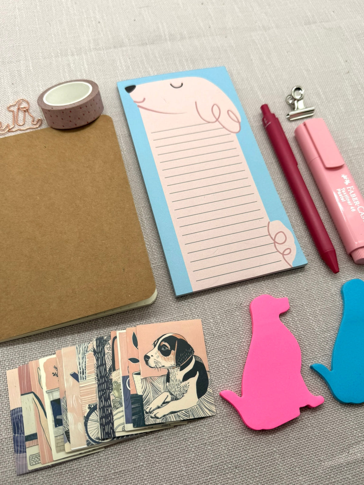 Ein Schreibset für die tägliche Organisation: Notizbuch, Notizblock mit Hundemotiv, daneben ein Kugelschreiber und ein Textmarker, beide in Pink. Unten liegen Haftnotizen in Form eines Hundes, sowie ein Stapel Sticker in Rosa und Blau. Oben sieht man Büroklammern in Hundeform, ein Washi-Tape und eine kleine Briefklemme